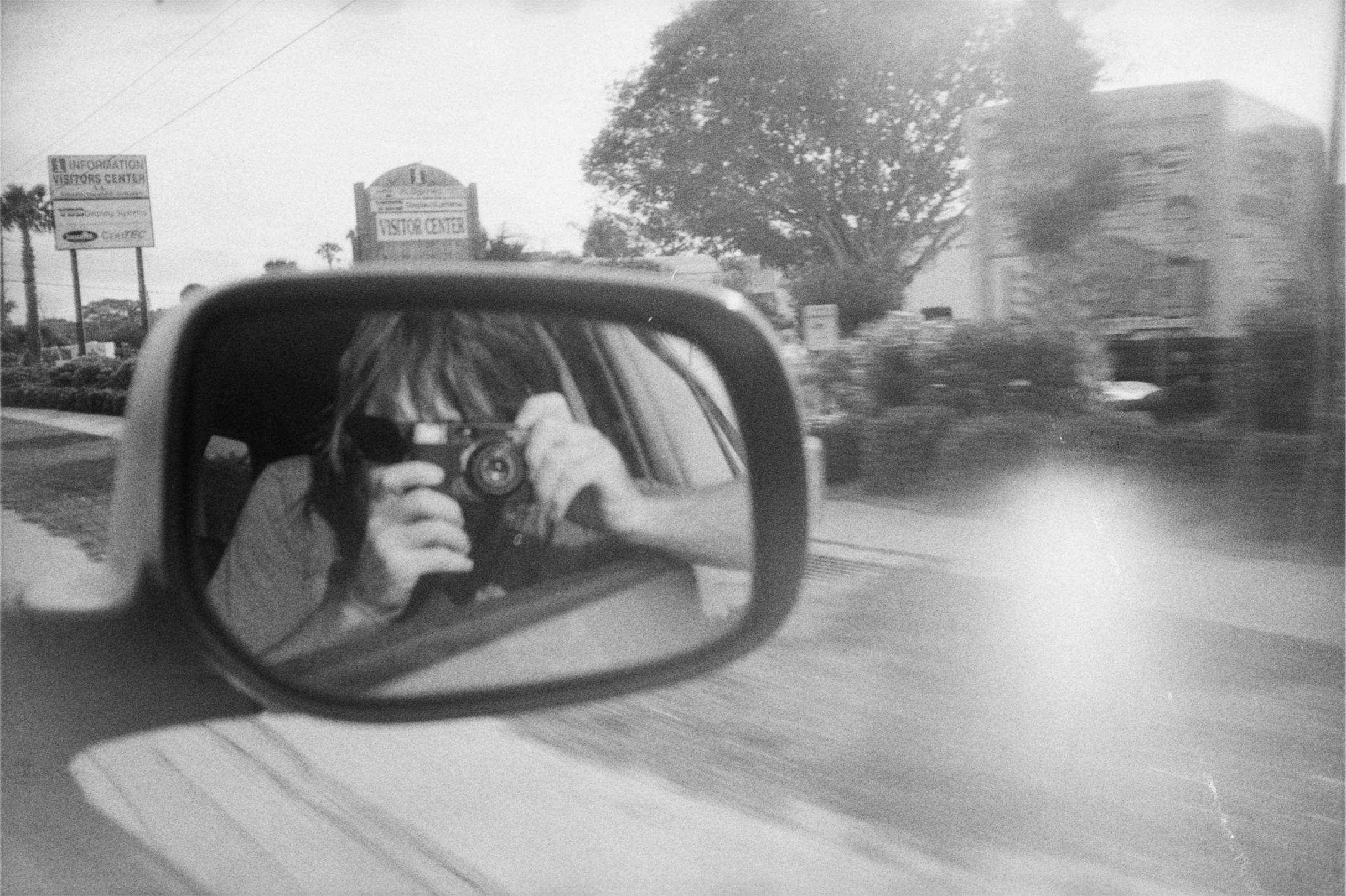 A hairy white man holding a film camera in the reflection of a car side mirror