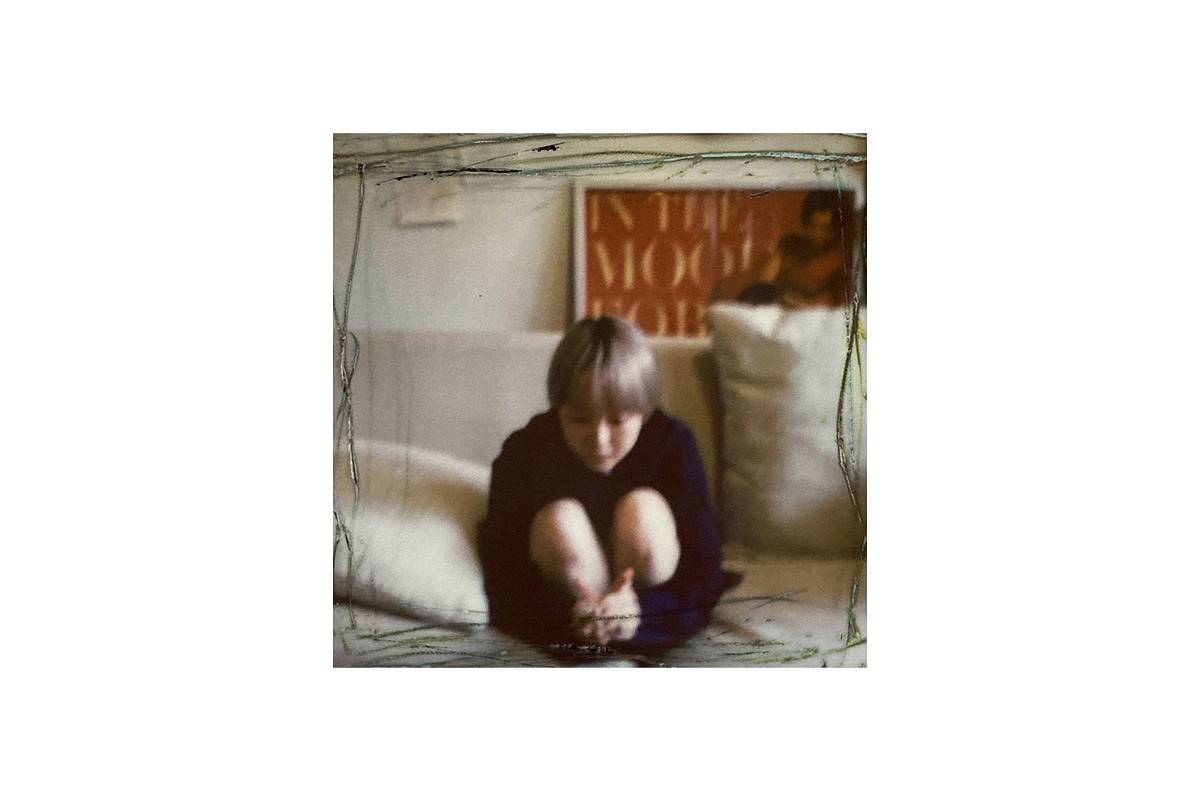 A candid colour photograph of a person sitting in a contemplative pose on a sofa, shot on instant film with a square format.