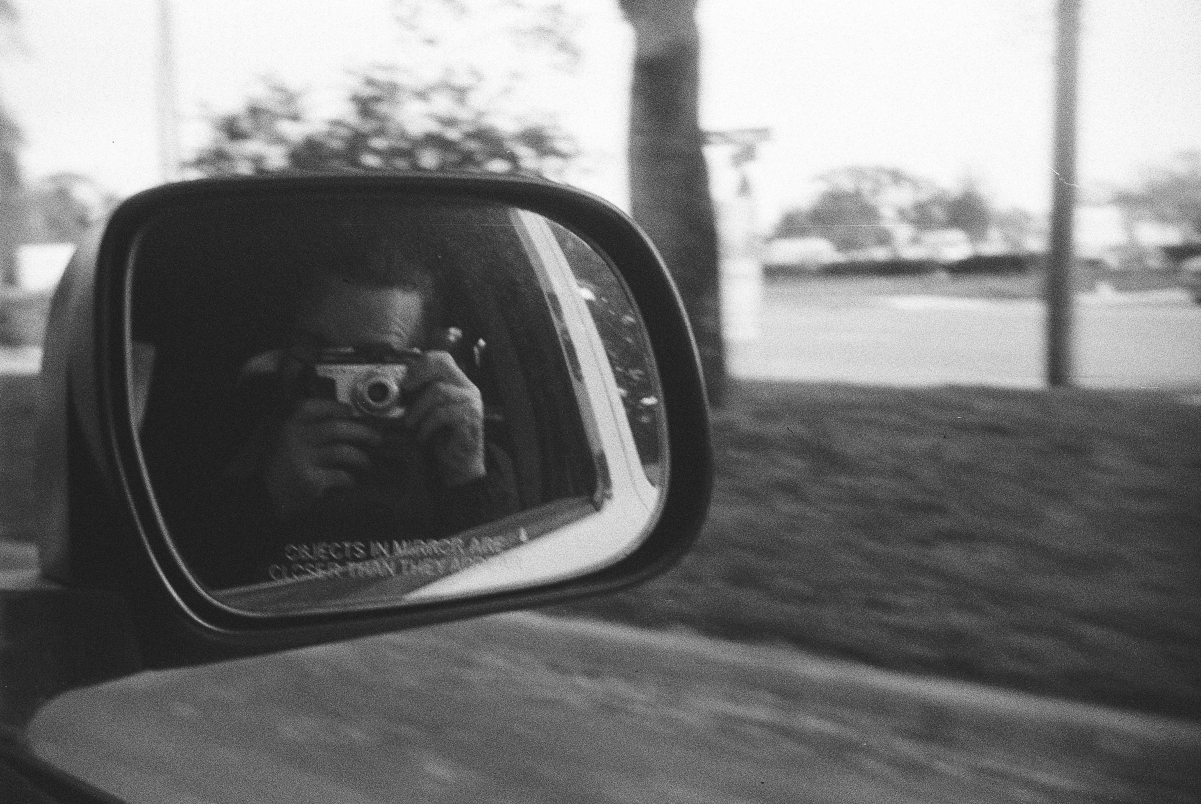 A hairy white man holding a film camera in the reflection of a car side mirror